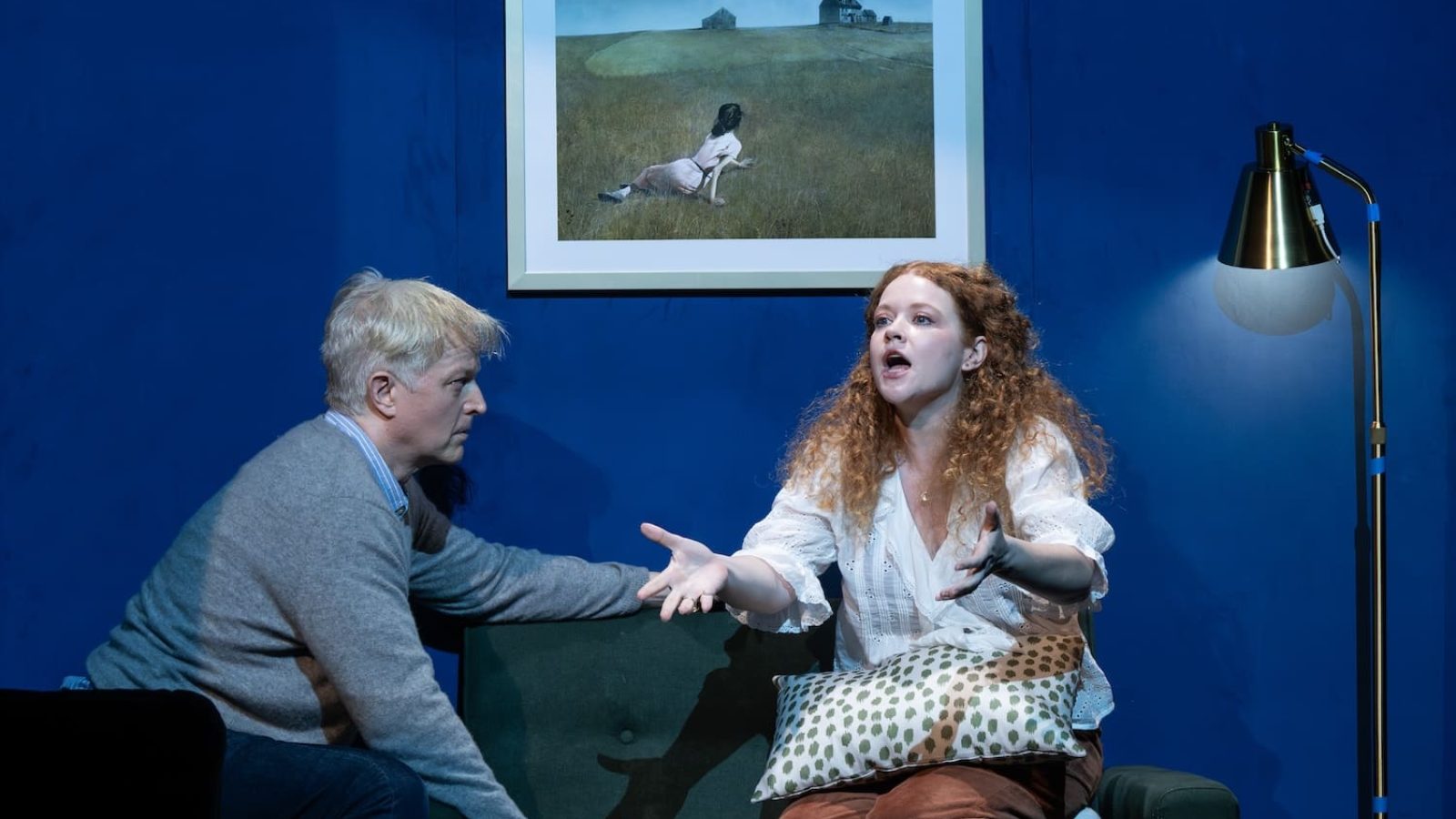 Scene from a play: woman gesturing emotionally to a man seated on a couch, under a framed picture.