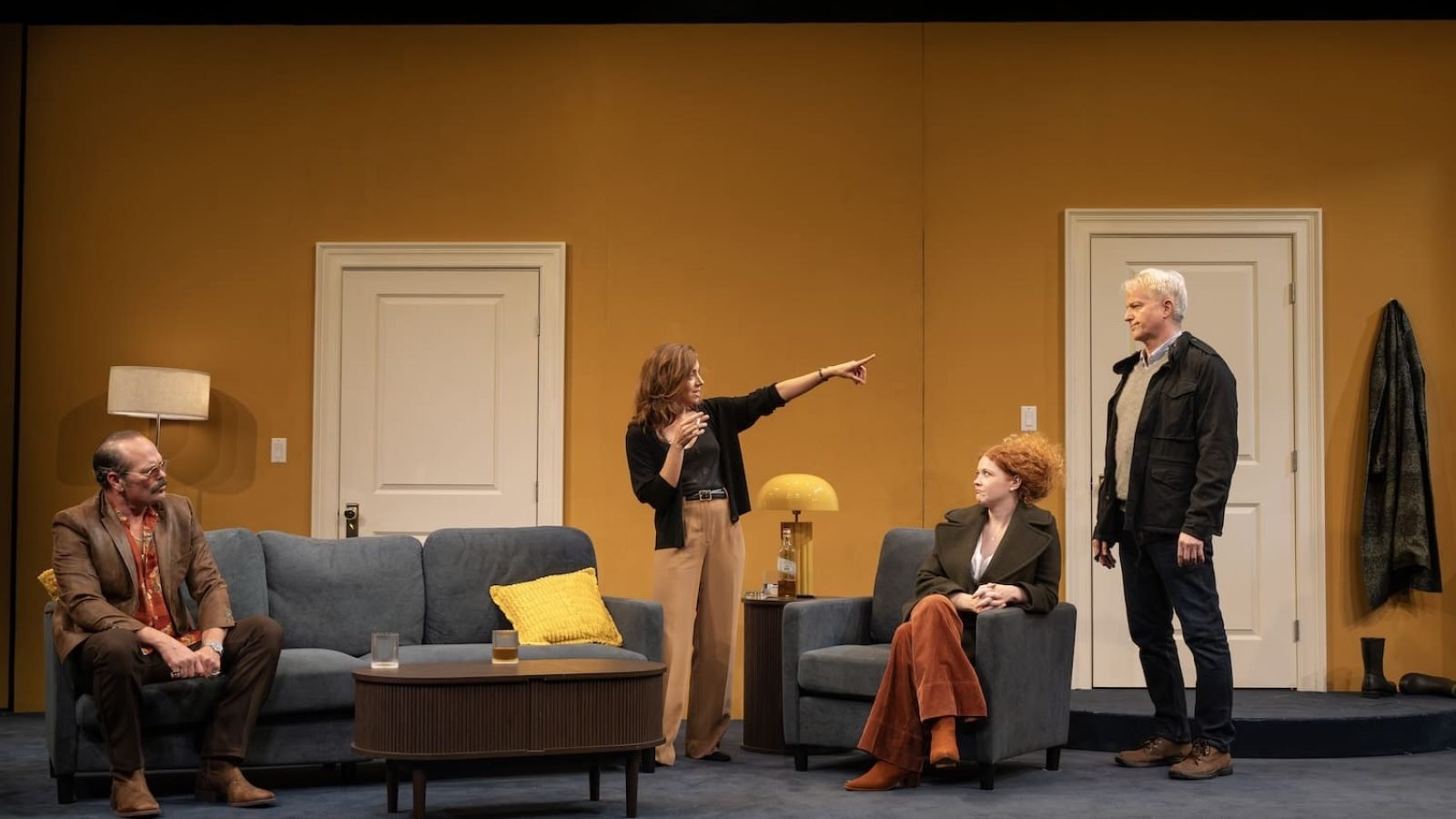 Theater scene: Four actors in a living room, one pointing. Stage production.