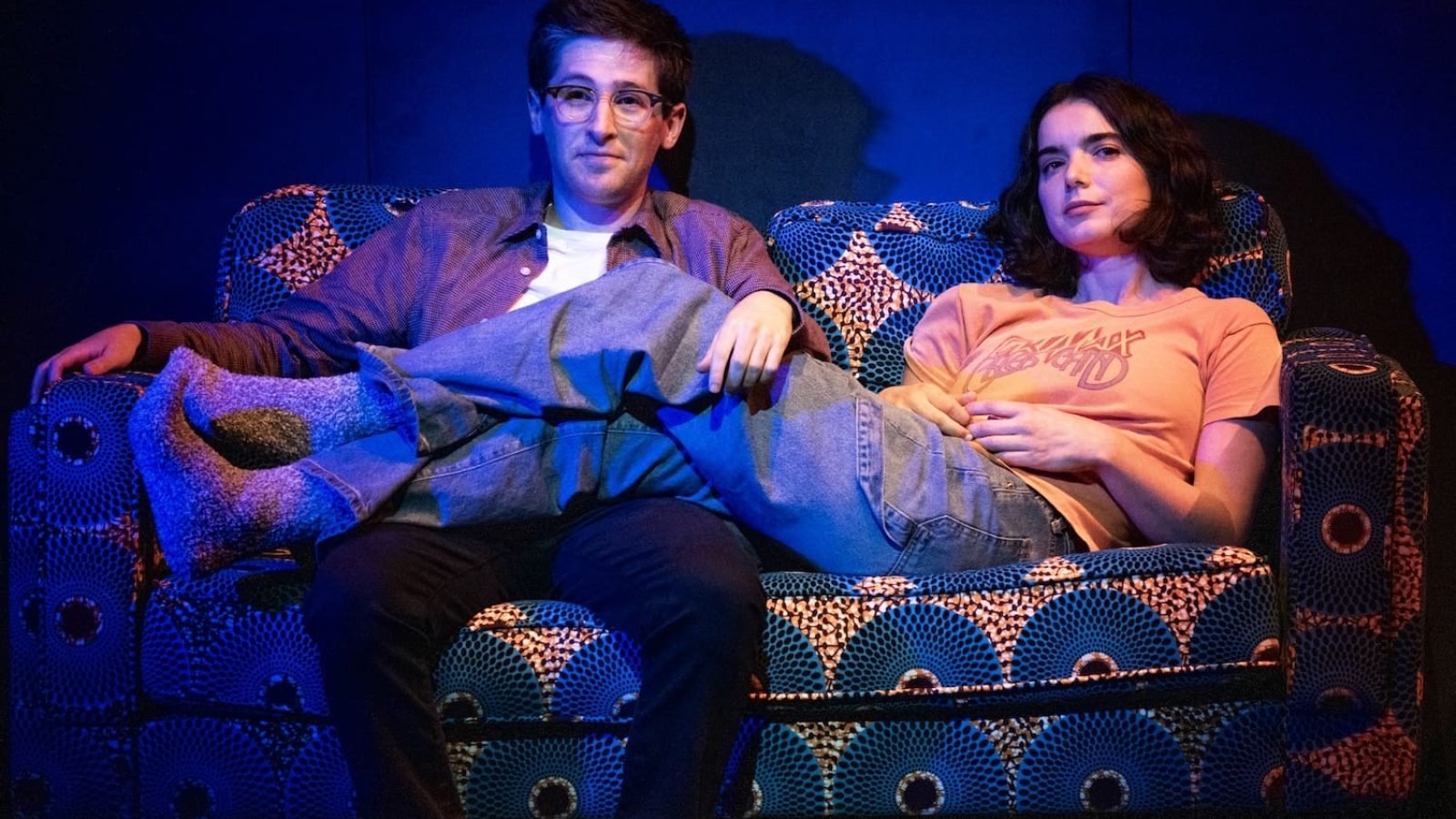 Two young people relaxing on a patterned couch under blue lighting.