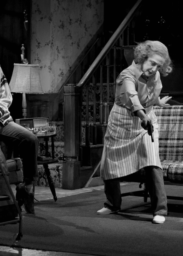 Bette Davis in a play, pointing a gun at another woman in a living room scene.