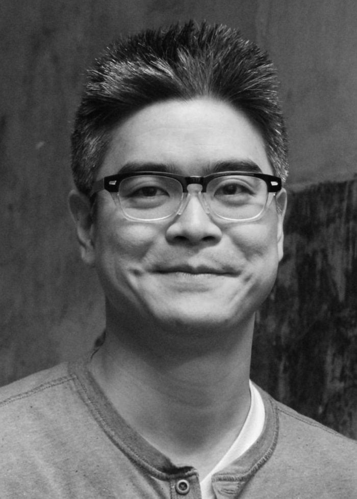 Lloyd Suh, wearing glasses, smiles for the camera in a black and white portrait.