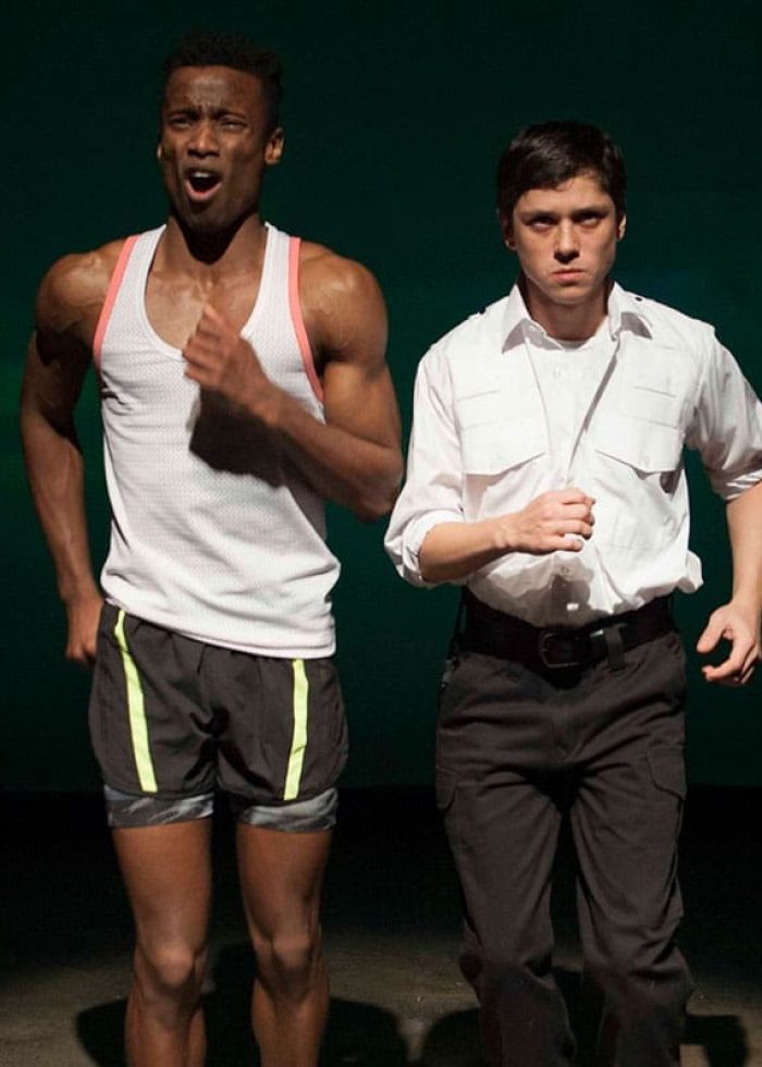 Two men running on stage. One in athletic wear, the other in a white shirt and dress pants.