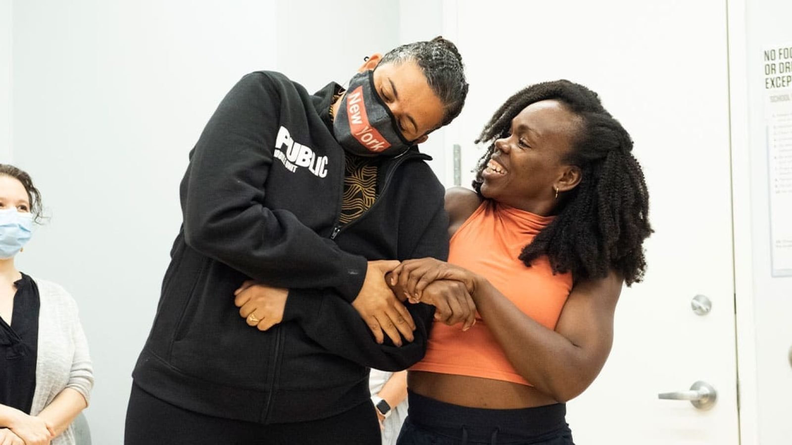 Two women embrace, one wearing a "New York" mask, during a rehearsal.