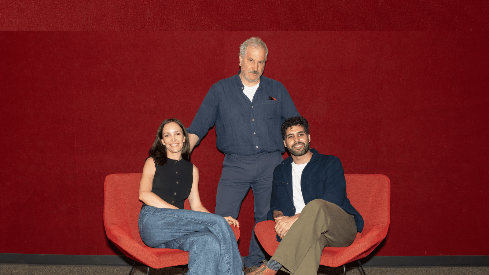 Three people sit in red chairs against a red wall. A man stands behind them.