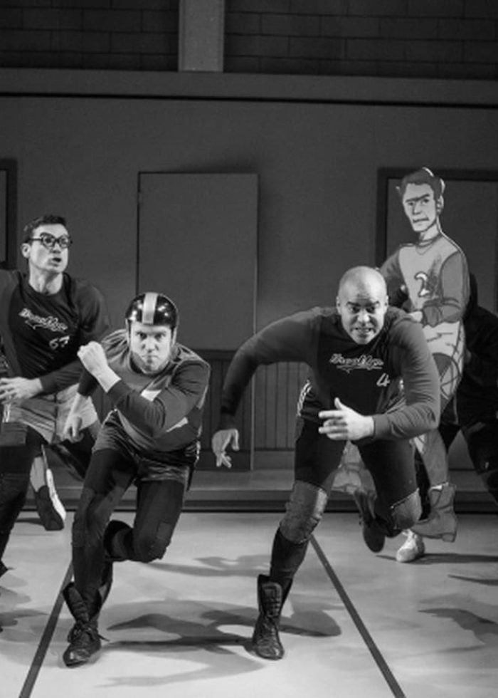 Cast of "The Jammer" running on stage, wearing jerseys and helmets. Dramatic black and white photo.