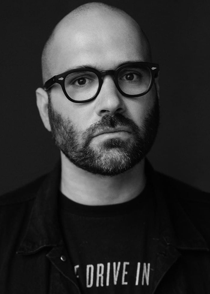 Marco Ramirez, bald man with beard and glasses, wearing a black shirt that says "DRIVE IN