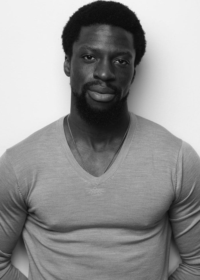 Michael Luwoye in a black and white portrait, wearing a gray v-neck sweater.
