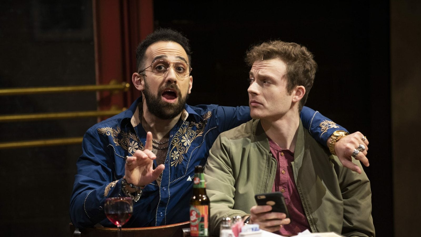 Two men sitting at a table, one wearing a blue shirt and the other a green jacket. They appear to be in a bar setting.