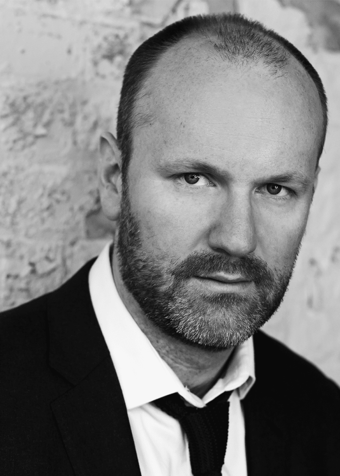 Andrew Musselman, headshot in black and white suit, leaning against a brick wall.
