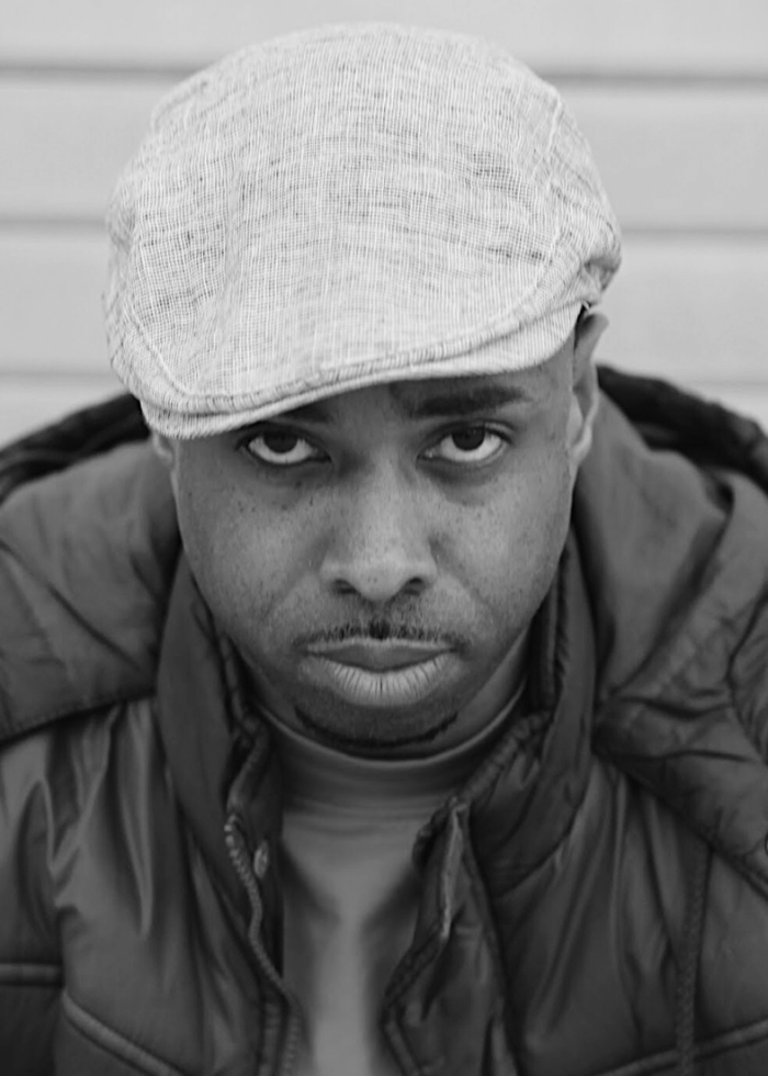 NSangou Njikam portrait in a newsboy cap and jacket, looking intensely at the camera.
