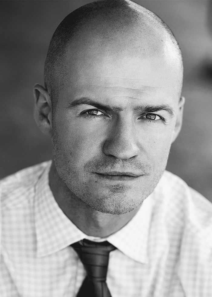 Close-up portrait of Adam O'Byrne, a man with a shaved head and a serious expression, wearing a dress shirt and tie.