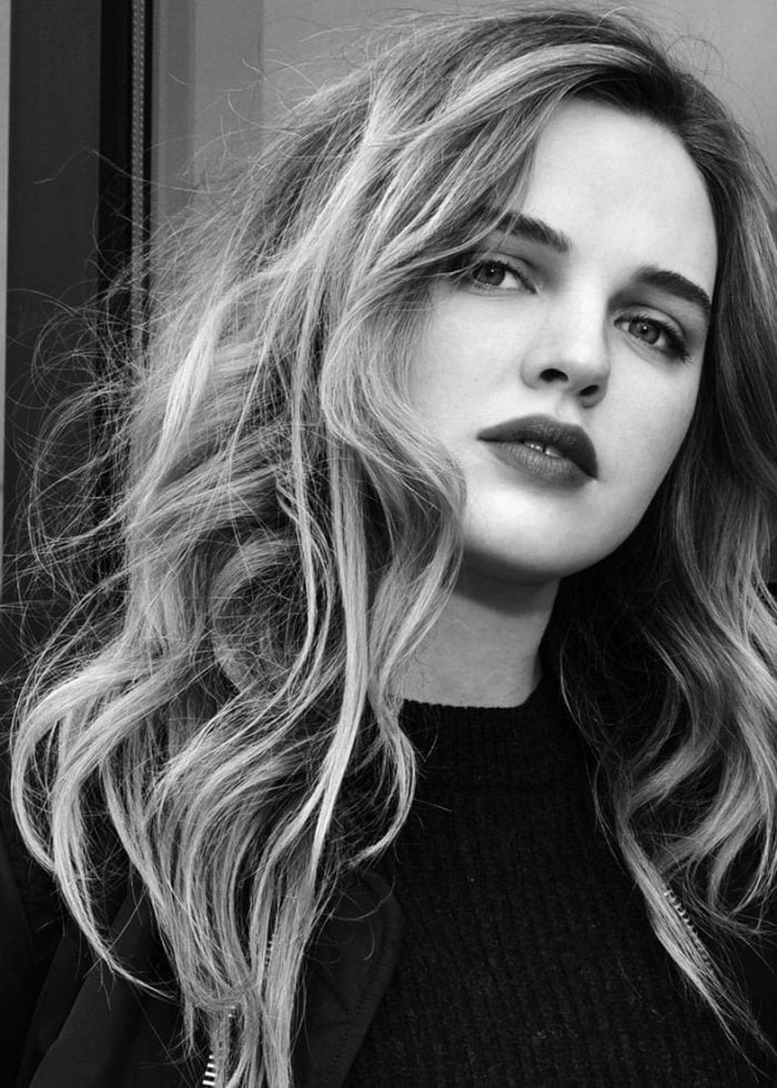 Odessa Young with wavy blonde hair and dark lipstick, looking at the camera.
