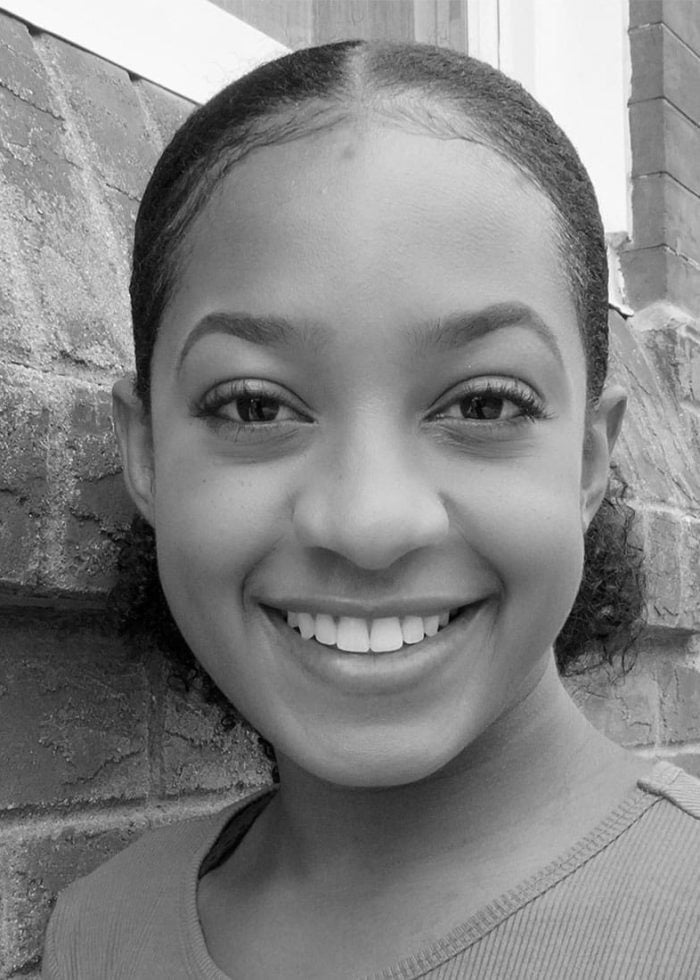 Smiling young woman with slicked-back hair, leaning against a brick wall.