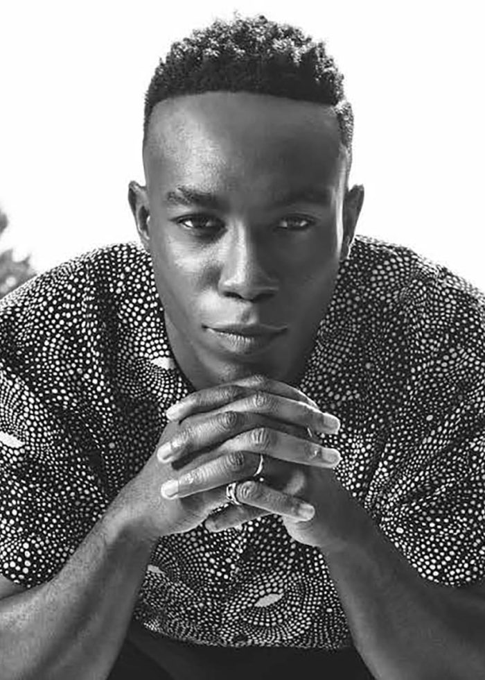 Olly-Sholotan-Headshot-bw Olly Sholotan poses with hands clasped, wearing patterned shirt