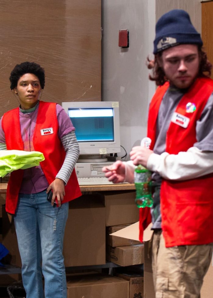 Two actors in red vests, possibly from a play titled 'Paris', stand inside a big box store setting. One holds a stack of shirts.