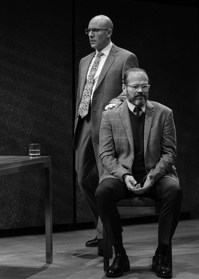Two men in suits on stage, one standing, one seated. Black and white photo.