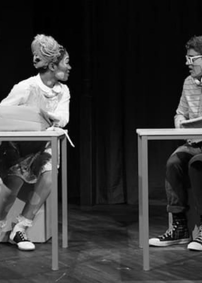 Actors in "Polkadots: The Cool Kids Musical" performing a scene, seated at desks.