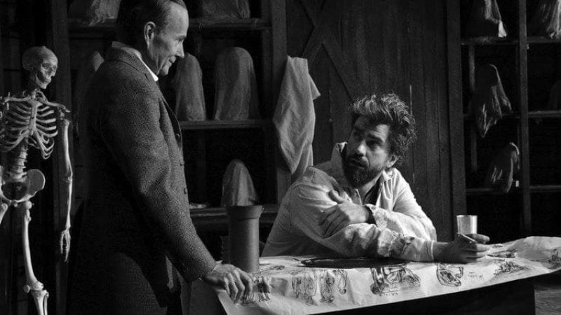 Two men in a black and white scene, one looking at the other. A skeleton and anatomical drawings are visible.