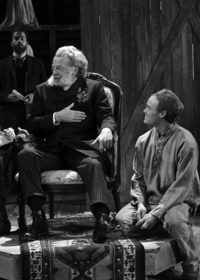 Black and white stage photo from 'Posterity': actors in period costumes. A man in a chair is the focus.