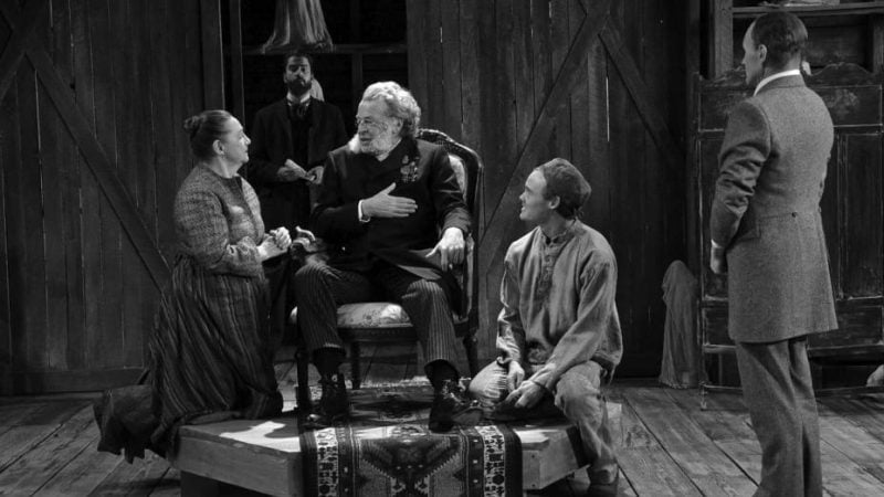 Black and white stage photo from 'Posterity': actors in period costumes. A man in a chair is the focus.