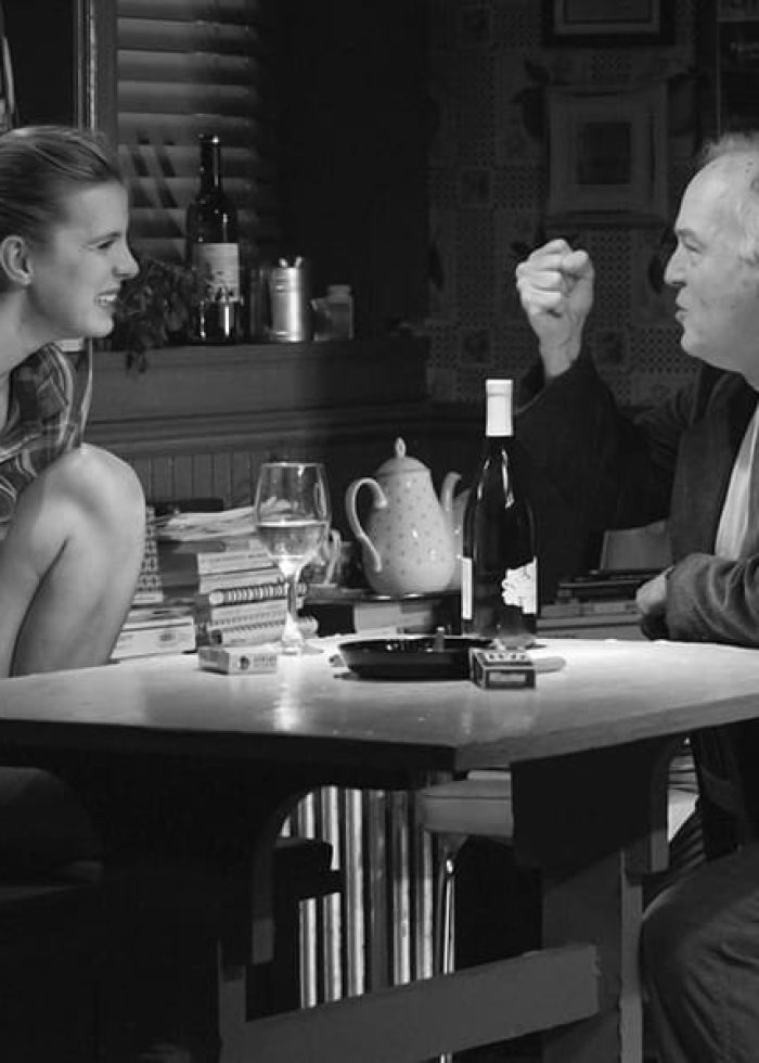 A man and woman in a dimly lit room, possibly from the play "I'm Gonna Pray for You So Hard," seated at a table with wine.