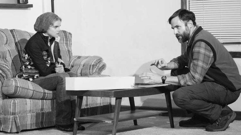 Man and woman in a black and white photo. Man crouches near a table, woman sits in an armchair.