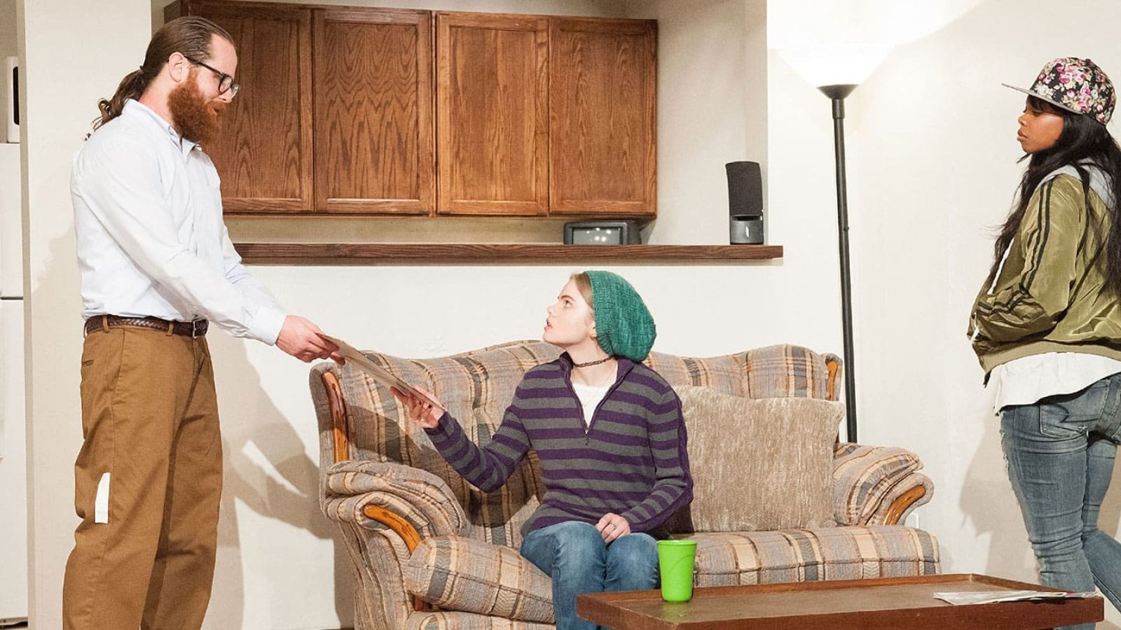 Three people in a living room; one hands papers to another while a third looks on.