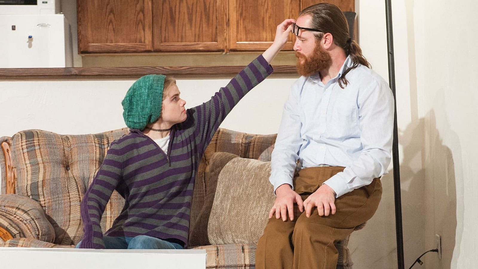 Man and woman sitting on couch, woman placing glasses on man's head.