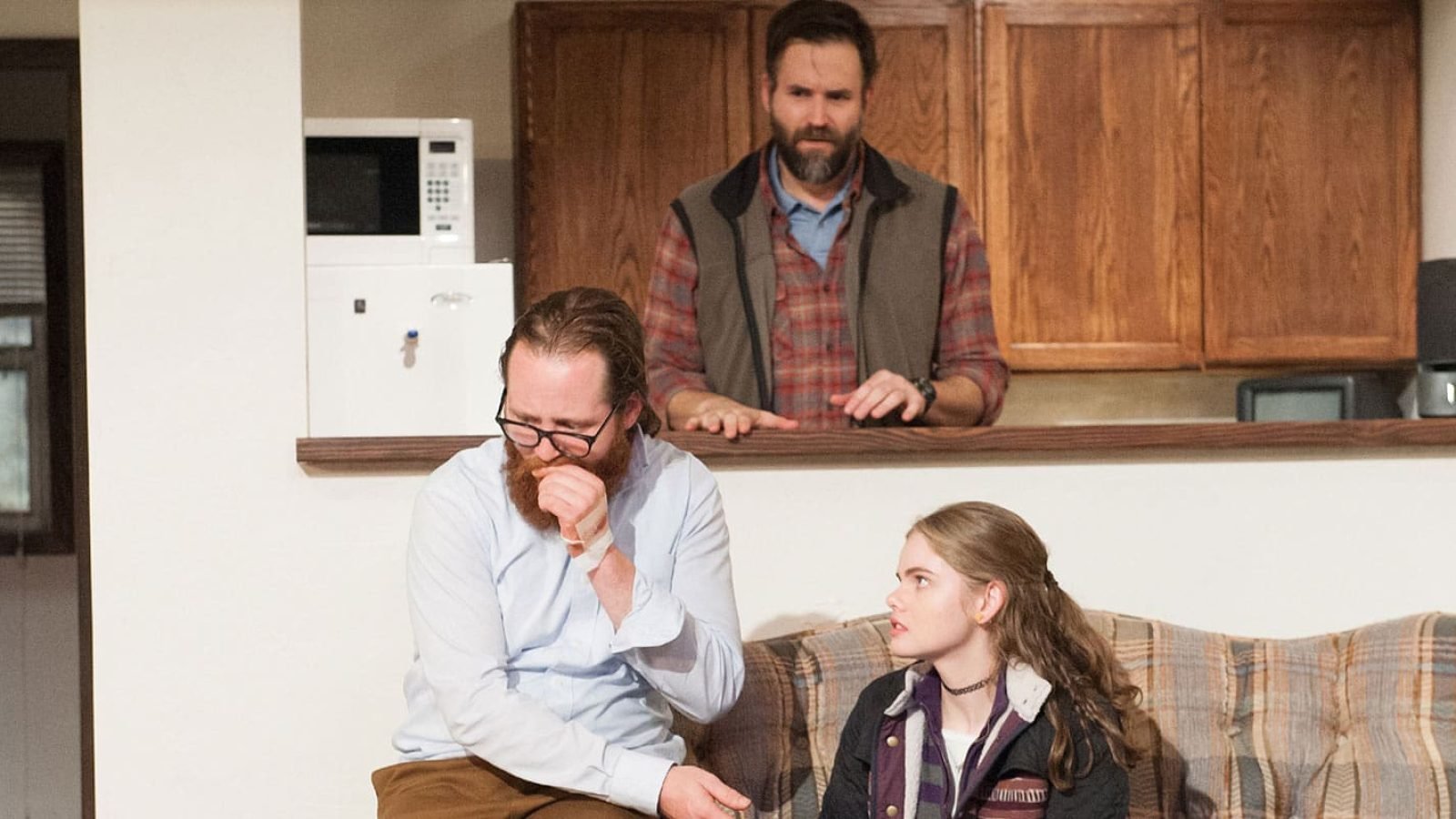 Three people in a living room, one man standing at a counter, two seated on a couch.