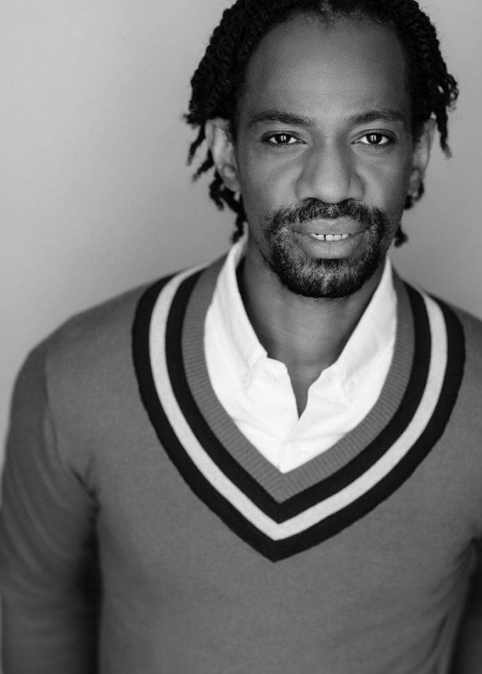 Reggie D. White, a man with dreadlocks, wearing a sweater and button-down shirt, looking at the camera.