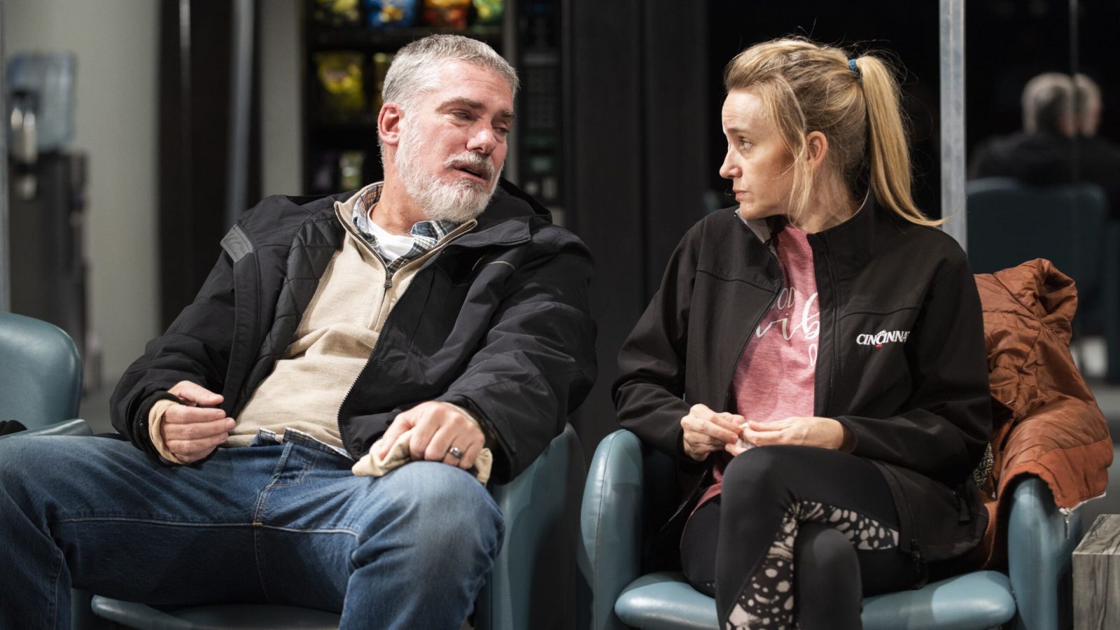 A man and woman sit in chairs, engaged in conversation. They appear to be in a waiting area.