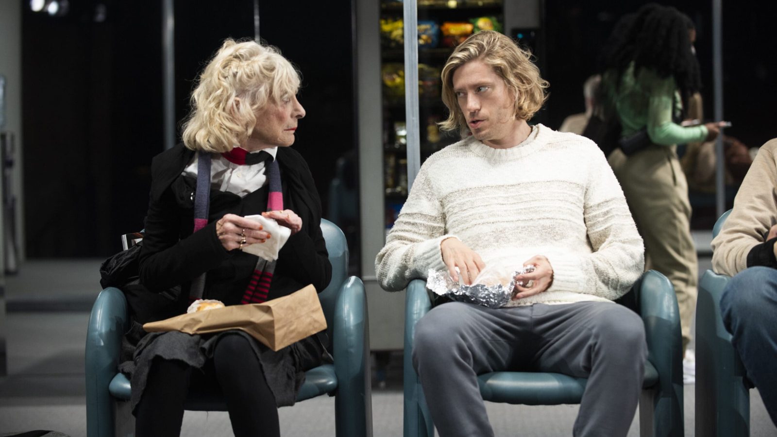Older woman and younger man converse while eating snacks in a waiting area.