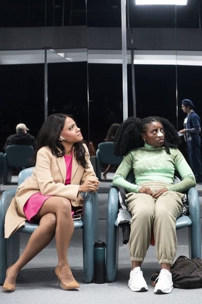 Two women sit in chairs in a waiting room, one wearing a beige coat and pink dress, the other in a green top and beige pants.
