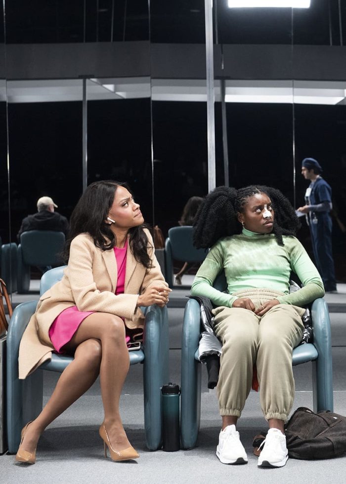 Two women sit in chairs in a waiting room, one wearing a beige coat and pink dress, the other in a green top and beige pants.