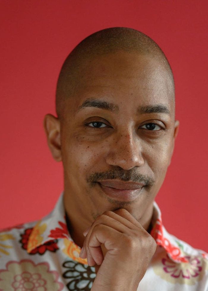 Ron S. McCan smiling, wearing a floral shirt, with hand on chin against a red background.