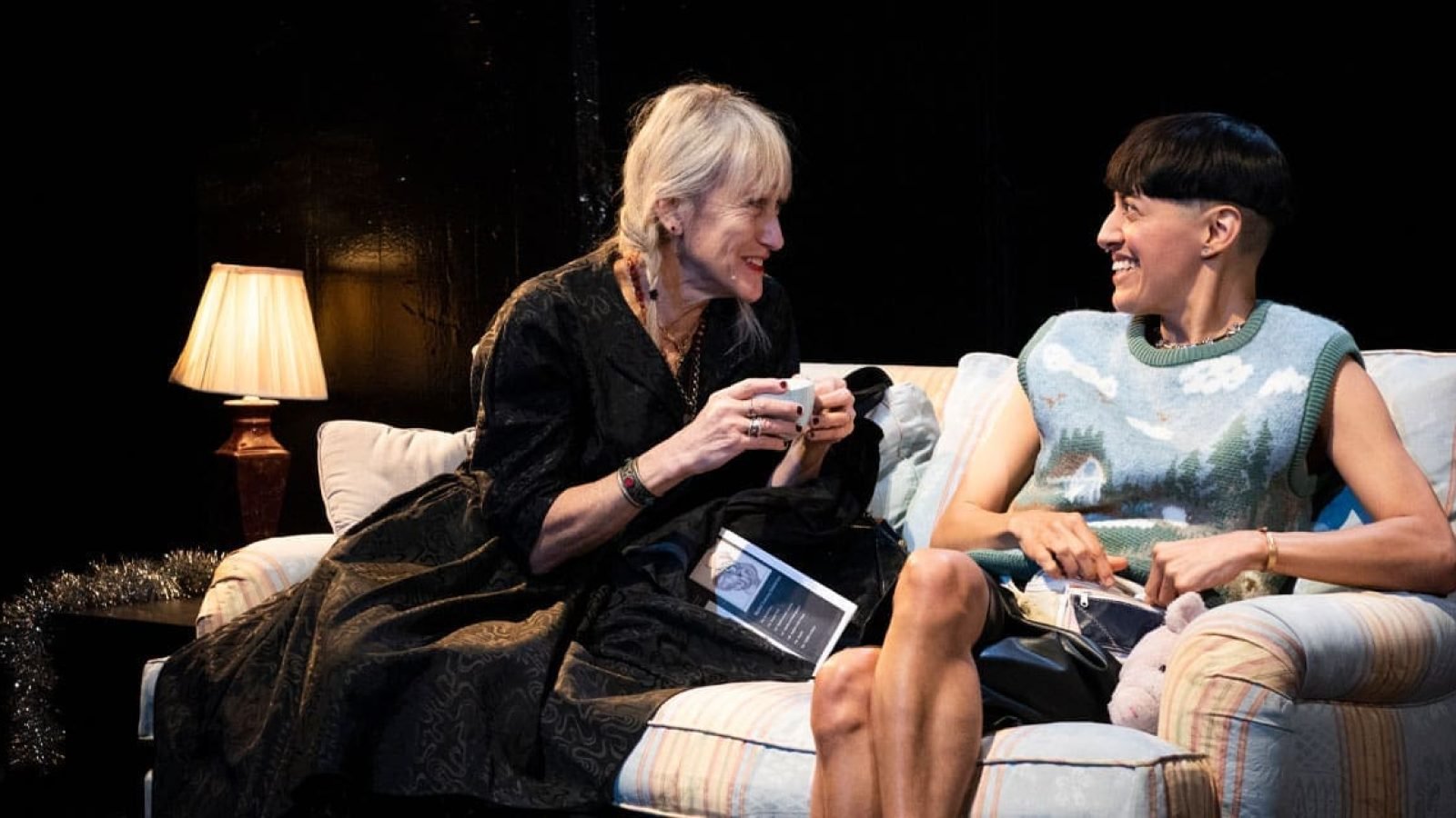 Two women share a quiet moment on a couch, enjoying tea and conversation.