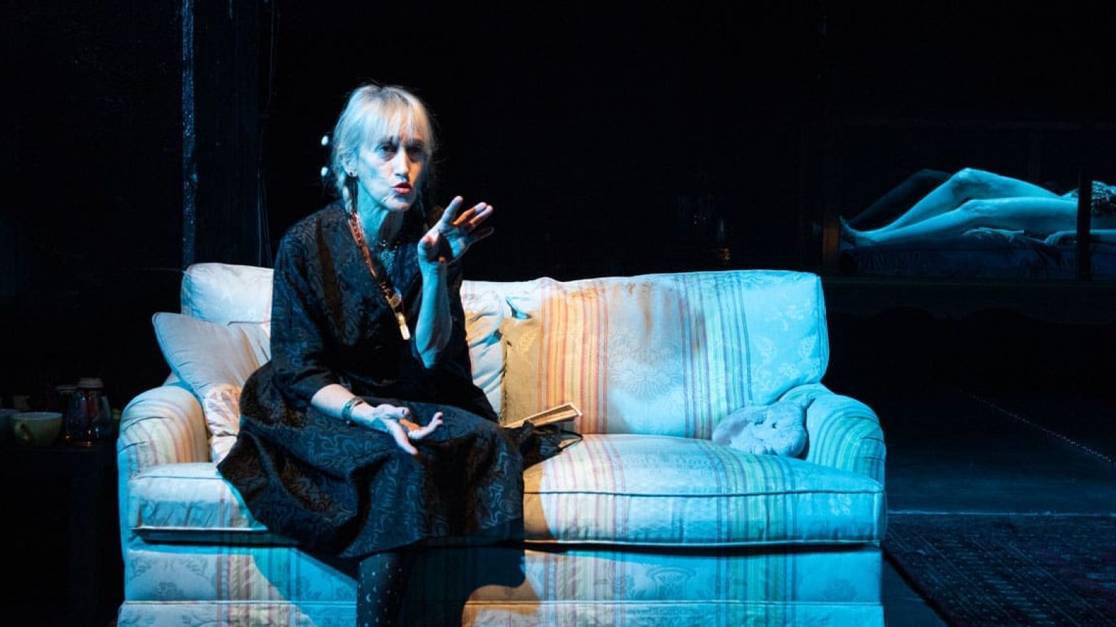 Woman in black dress sits on a couch, gesturing; a figure lies in a glass enclosure in the background. Shhh.