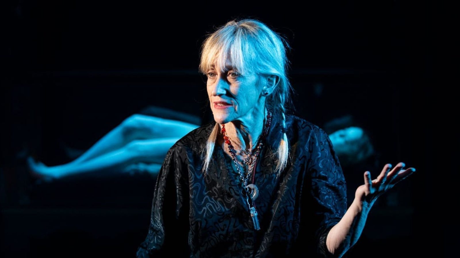 A woman with blonde hair in a dark dress gestures, a blurry figure lying down behind her. Shhh.