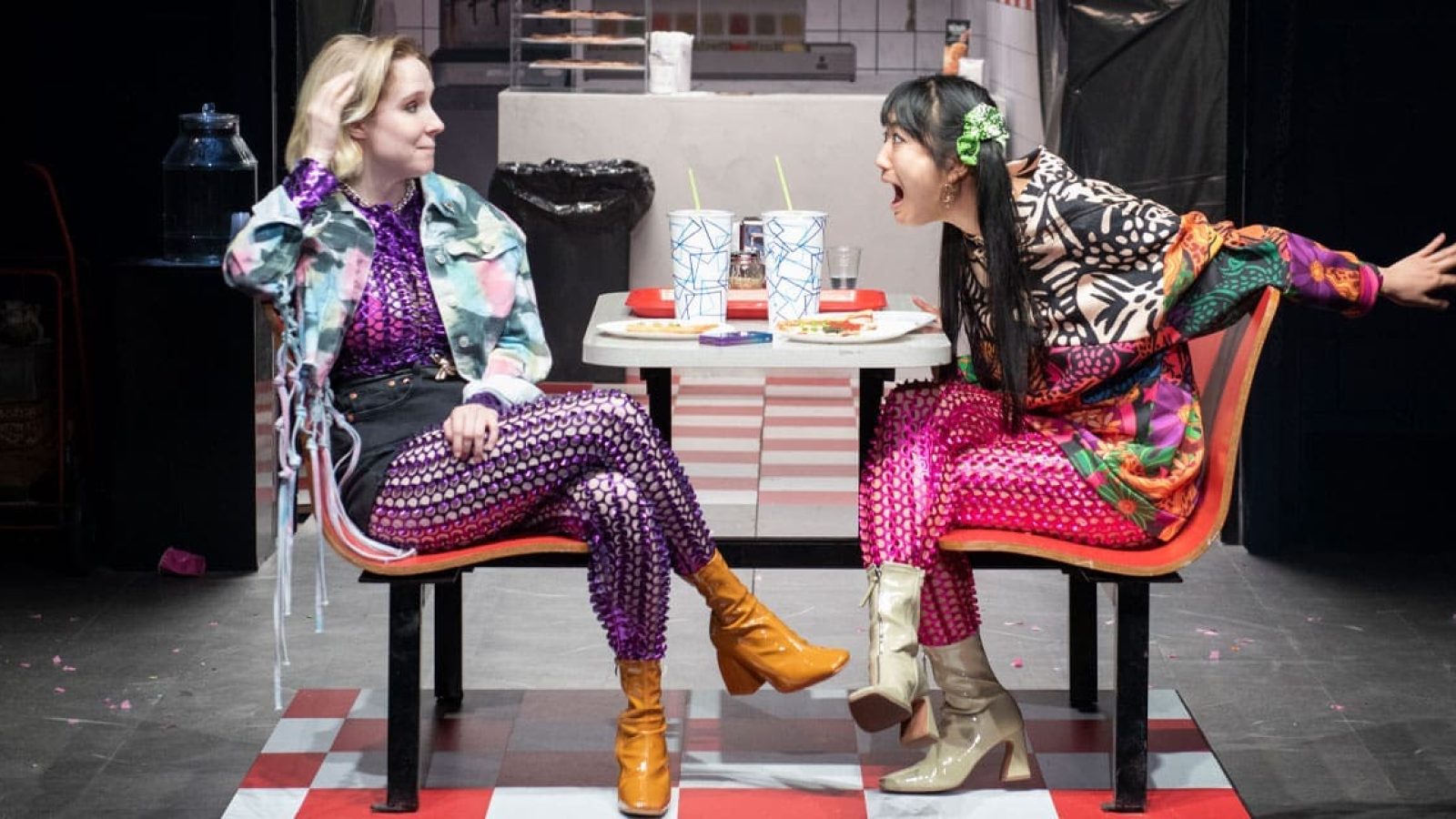 Two women in colorful outfits sit at a diner booth, one whispering, creating a hushed atmosphere.