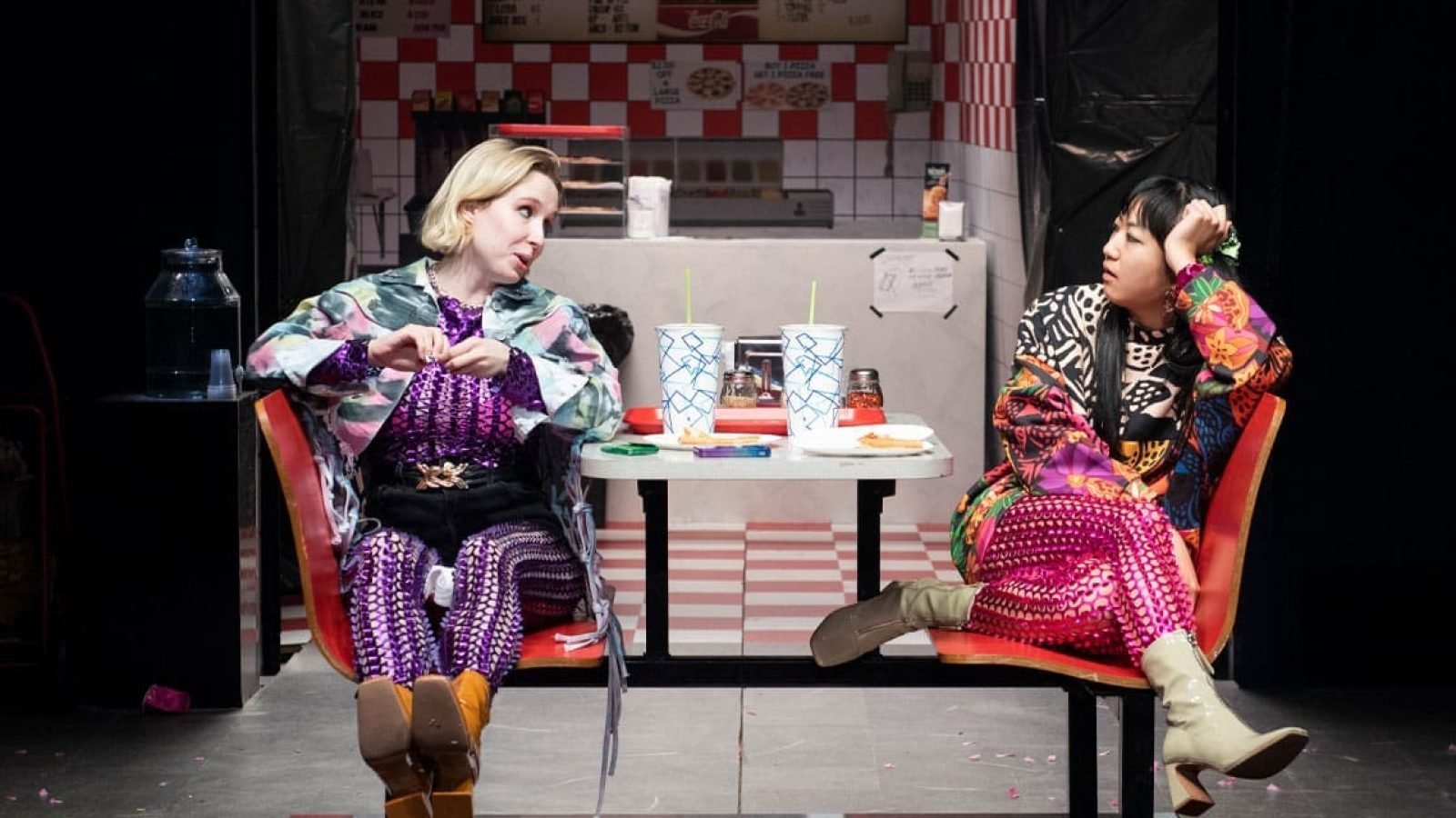 Two women in vibrant outfits sit at a diner booth, engaged in quiet conversation. Shhh.