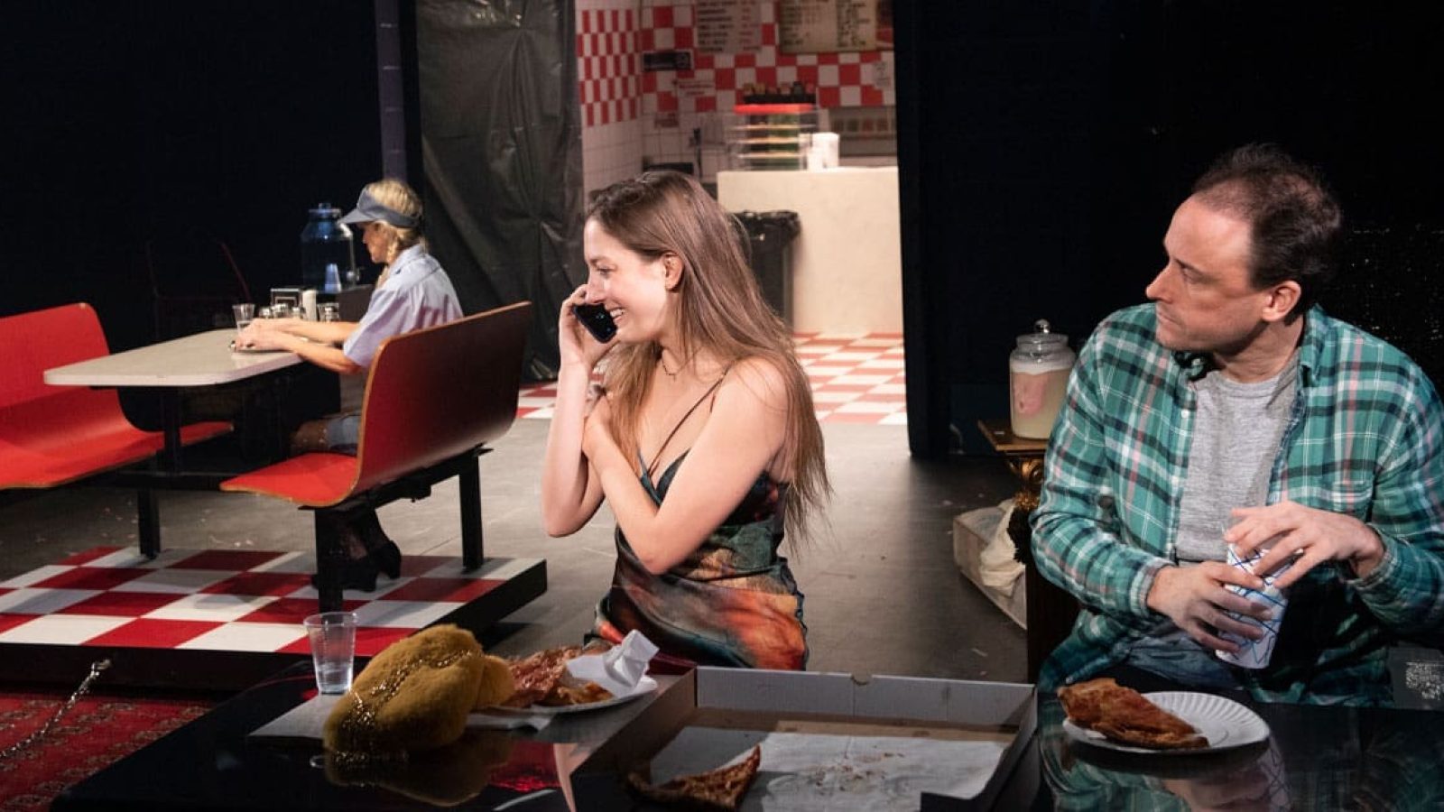 Three people sit in a diner booth, one woman on a phone call, another seated at a nearby table, and a man eating pizza.