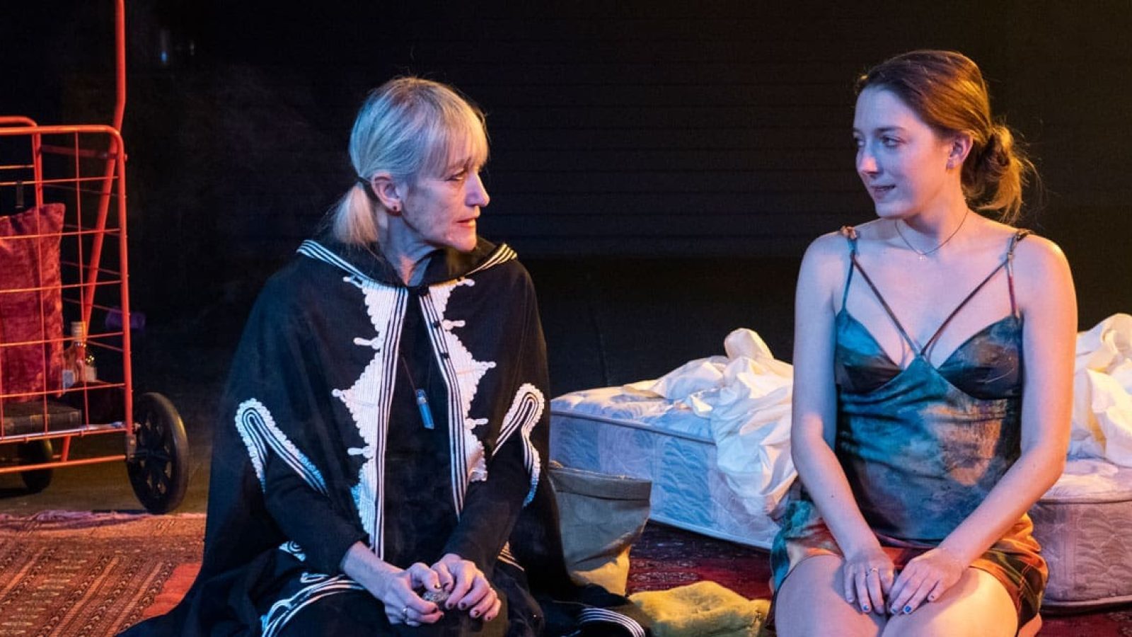 Two women sit on a rug, one older in a dark cape, the other younger in a slip dress, near a mattress. Shhh.