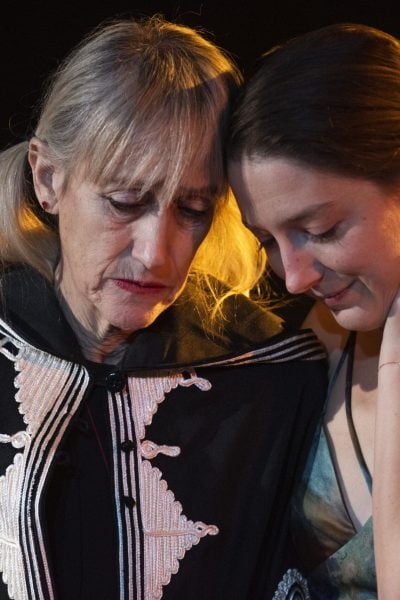 Two women embrace, heads together, whispering a secret. One wears a dark cape with ornate detailing.