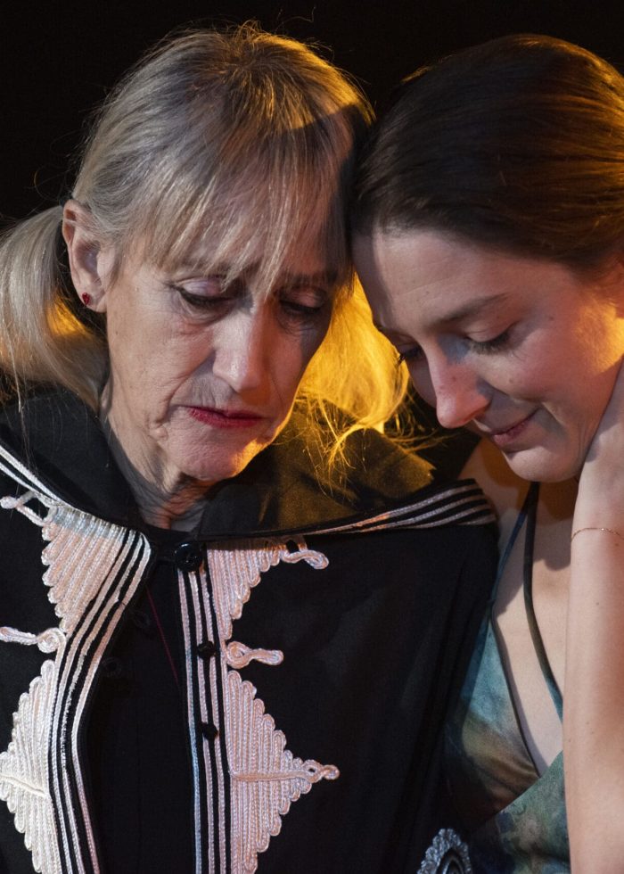 Two women embrace, heads together, whispering a secret. One wears a dark cape with ornate detailing.