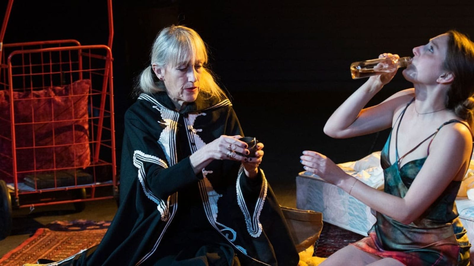 Two women, one in a dark cloak, the other in a slip, share a quiet moment. One drinks from a bottle.