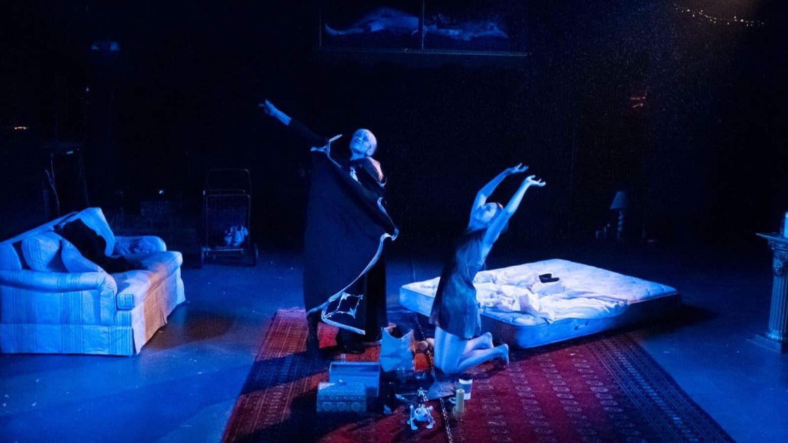 Two performers in a dimly lit theater, one in a long robe, the other kneeling on a mattress. Shhh.