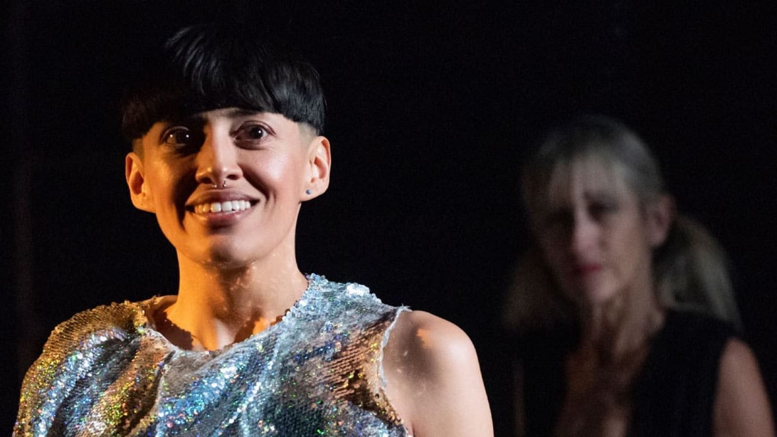 Close-up of a woman with short, dark hair, smiling. She is wearing a sparkly silver top.