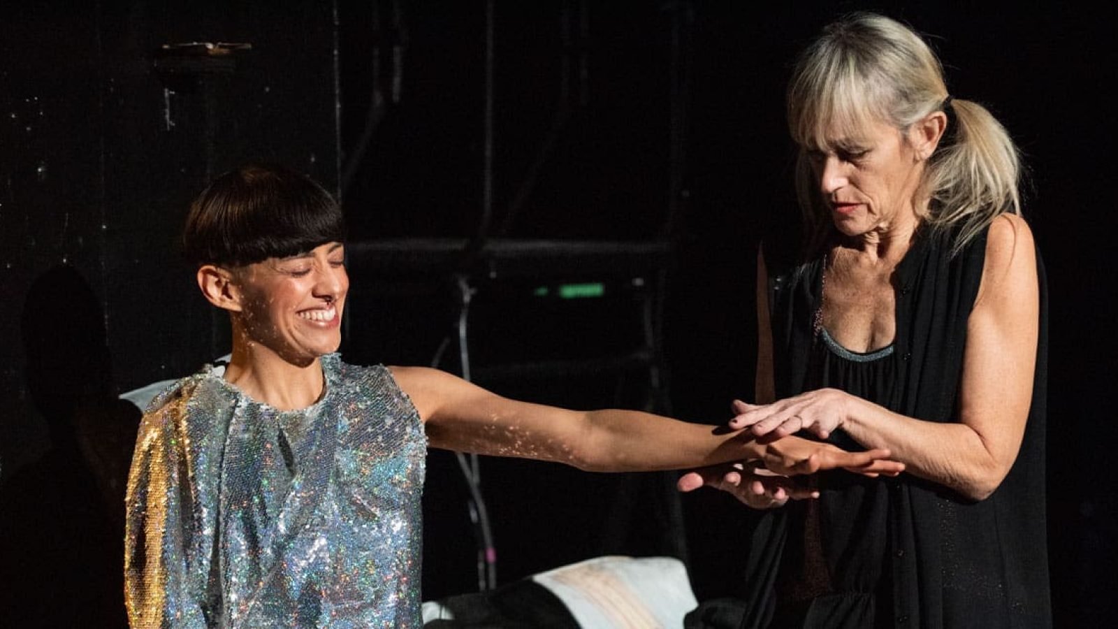 Two women, one in a sequined top, interact; a moment of quiet connection.