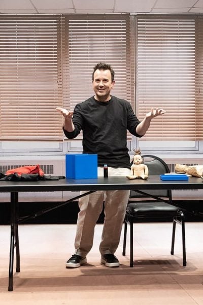 Man in black shirt presenting at a table with a blue cube and doll, resembling a simulacrum.