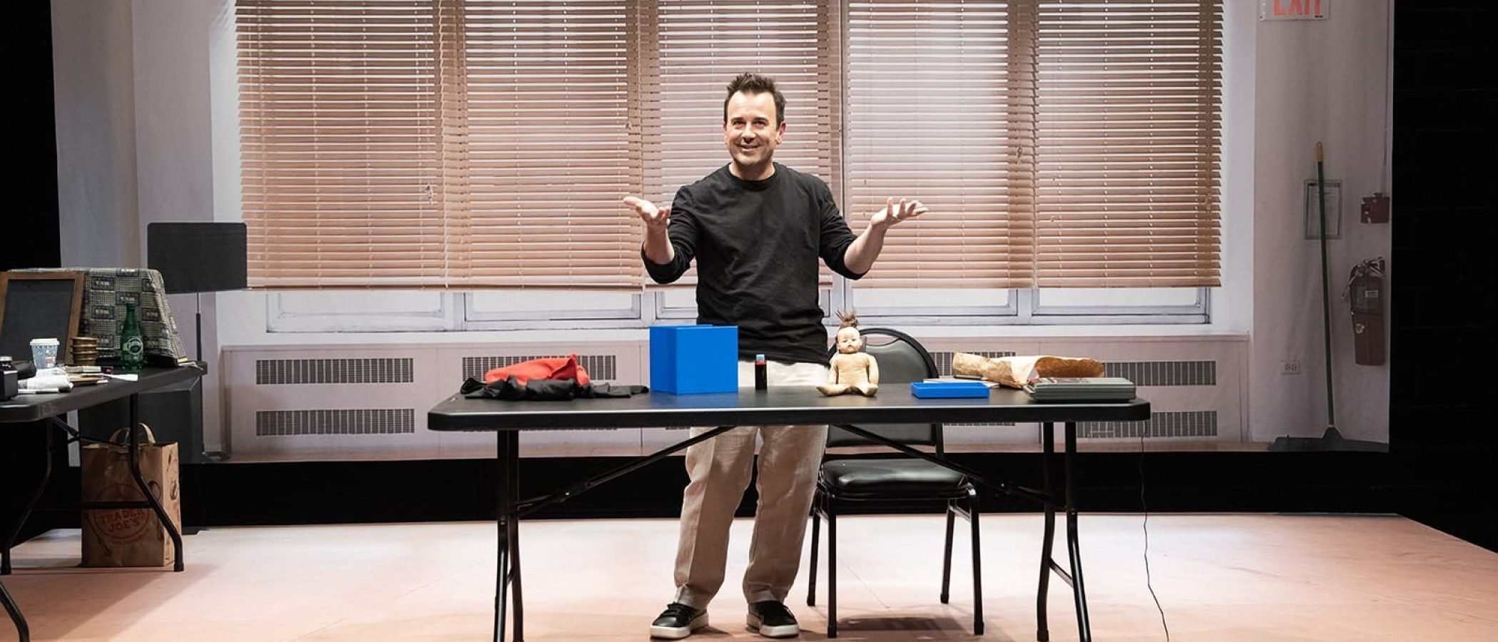 Man in black shirt presenting at a table with a blue cube and doll, resembling a simulacrum.